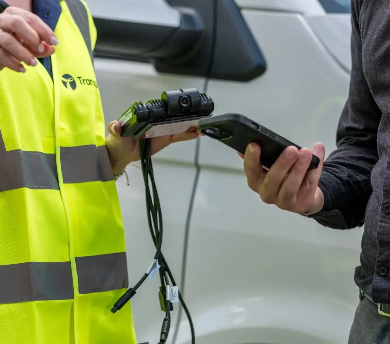 Transpoco employee in a high-vis jacket and holding an AI-connected camera, standing next to a van beside a driver who's looking at data on his smartphone