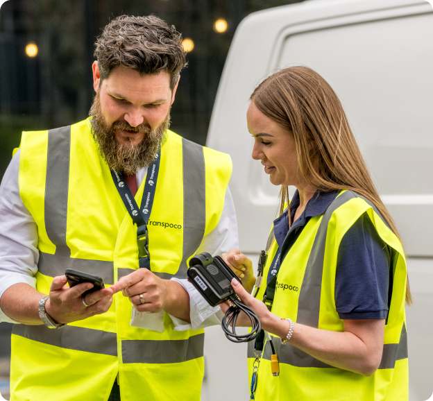 Two Transpoco employees in high-vis jackets standing next to a white van, looking at data on a smartphone