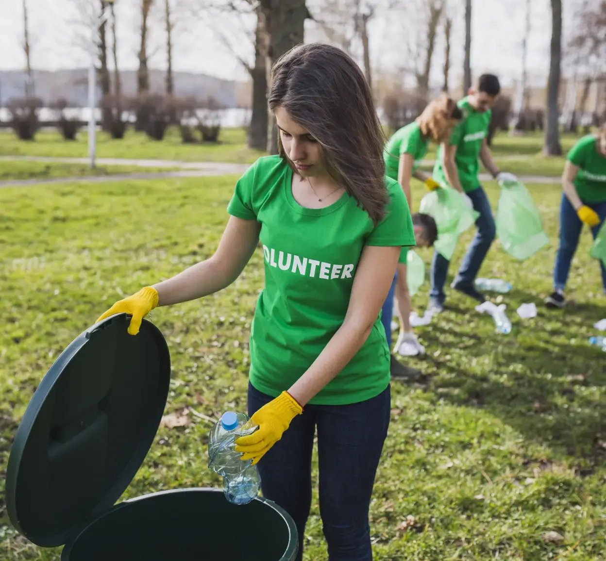 Volunteer in a green shirt and yellow gloves standing in a field, putting plastic litter in a black bin