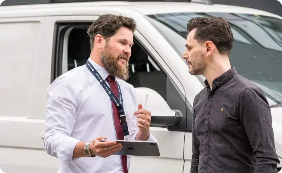 A Transpoco employee in a light purple shirt standing in front of a van, holding a tablet and talking to a driver