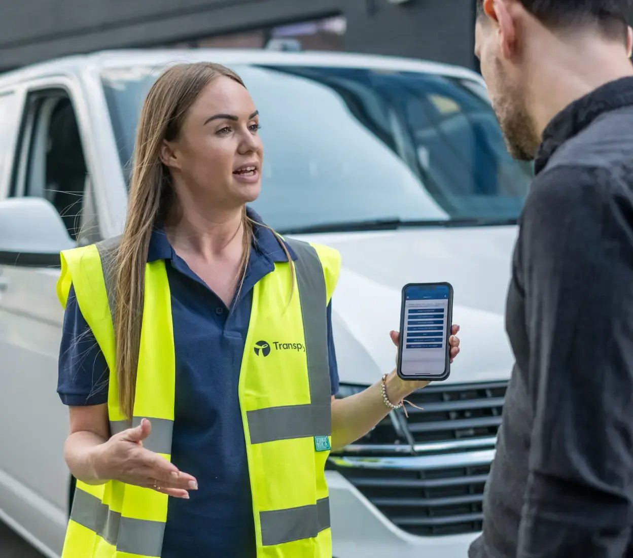 Transpoco employee in a high-vis jacket standing in front of a van, showing vehicle telematics data to a driver on a smartphone